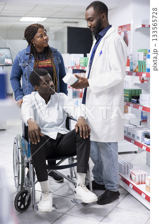 Pharmacist counseling a customer with disability on treatment options, discussing about medication dosage to support patient health. Professional medical staff showing drugstore pills. Pharmacist counseling a customer with disability on treatment options, discussing about medication dosage to support patient health. Professional medical staff showing drugstore pills. 133565728
