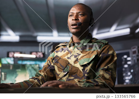 Military dispatcher in base of operations ready to respond to emergencies and communicate with soldiers on front lines. African american telecommunications operator in uniform talking in headset mic Military dispatcher in base of operations ready to respond to emergencies and communicate with soldiers on front lines. African american telecommunications operator in uniform talking in headset mic 133565766