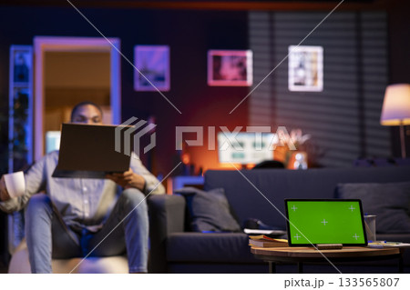 Black young casual man having green screen in his trendy apartment, stylish living room decorated with furniture and neon lights. Casual person relaxing on a bean bag at home, teleworking. Black young casual man having green screen in his trendy apartment, stylish living room decorated with furniture and neon lights. Casual person relaxing on a bean bag at home, teleworking. 133565807