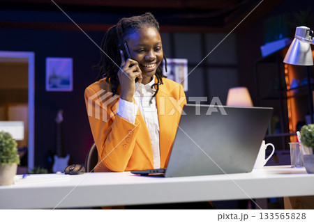 Cheerful teleworker talking in phone call with coworker, using laptop in apartment office to solve work tasks. Smiling teenager doing telephone call with colleague, working together to finish project 133565828