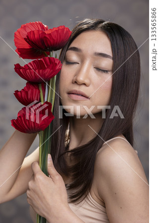 Beautiful asian woman model closing her eyes next to red carnations bouquet, posing with glossy smooth skin for a beauty skincare advertisement. Proud feminine pose with natural look. 133565866