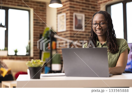 African american woman smiling brightly at device screen, reading online articles in brick wall apartment. Portrait of black female remote worker happily using laptop at desk, checking office emails. African american woman smiling brightly at device screen, reading online articles in brick wall apartment. Portrait of black female remote worker happily using laptop at desk, checking office emails. 133565888