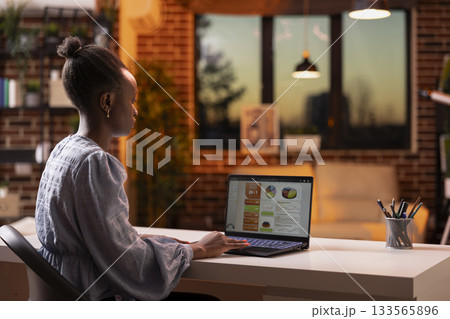 Modern black woman working remotely on business reports and presentations at desk. Confident female entrepreneur using laptop, focusing on online tasks and analyzing company sales revenue at home. Modern black woman working remotely on business reports and presentations at desk. Confident female entrepreneur using laptop, focusing on online tasks and analyzing company sales revenue at home. 133565896
