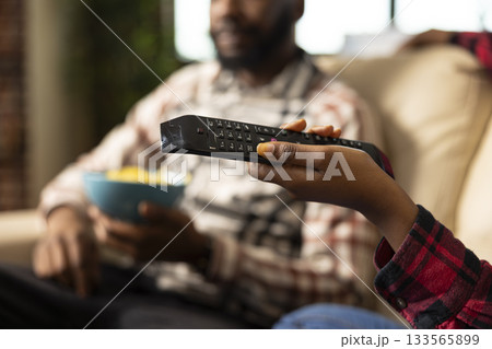 Close up on hand of black woman holding remote control, aimed at television as background shows man holding bowl of snacks. African american person using device to flip through tv channels. Close up on hand of black woman holding remote control, aimed at television as background shows man holding bowl of snacks. African american person using device to flip through tv channels. 133565899