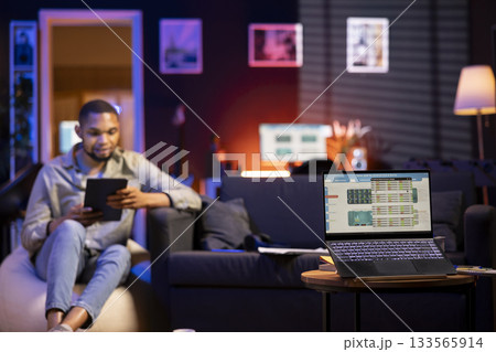 Young Black man relaxes on sofa and uses PC to track forex trades, analyzing stock market graphs for financial growth. Remote worker reviews income trends and trading data for online investments. Young Black man relaxes on sofa and uses PC to track forex trades, analyzing stock market graphs for financial growth. Remote worker reviews income trends and trading data for online investments. 133565914