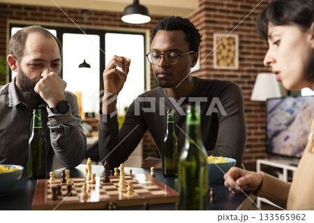 Multiethnic group of friends seated around table, enjoying a board game with snacks. African american man with chess piece in hand, looking at chessboard on table along with his caucasian companions. Multiethnic group of friends seated around table, enjoying a board game with snacks. African american man with chess piece in hand, looking at chessboard on table along with his caucasian companions. 133565962