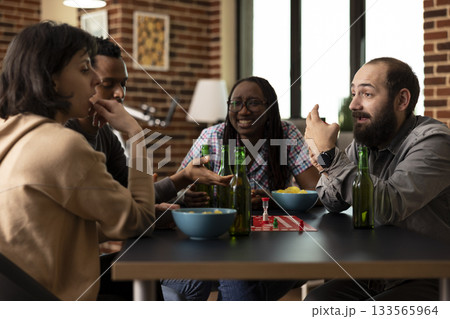 Young adults pause from board game to listen intently as white man shares an engaging story. Diverse friends gathered around the table with alcoholic beverage and bowl of chips, hanging out indoors. Young adults pause from board game to listen intently as white man shares an engaging story. Diverse friends gathered around the table with alcoholic beverage and bowl of chips, hanging out indoors. 133565964