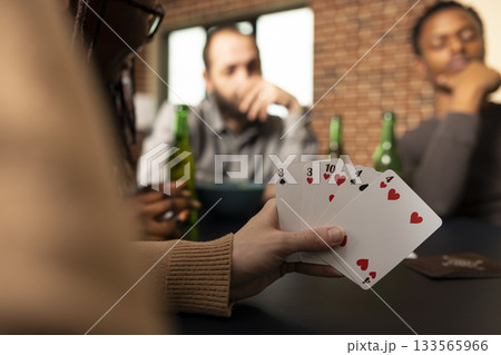 Selective focus on caucasian woman grasping poker cards during game day with diverse friends at home. Multiethnic group sits around living room table enjoying fun leisure activity in brick wall room. Selective focus on caucasian woman grasping poker cards during game day with diverse friends at home. Multiethnic group sits around living room table enjoying fun leisure activity in brick wall room. 133565966