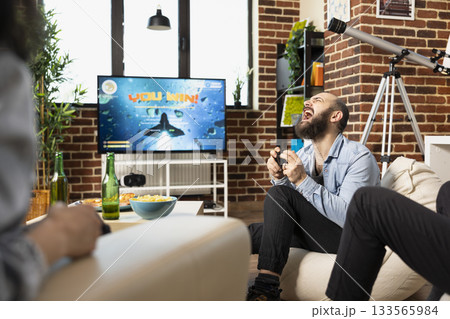 Caucasian man holds gamepad, shouting as he celebrates winning tough gaming tournament during group indoor party. Bearded male gamer filled with excitement after video game victory in apartment. 133565984