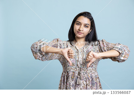 Dissatisfied upset girl acting displeased with a dislike symbol in studio, refusing something and giving a thumbs down against blue background. Young woman being negative, disagreement. 133566087
