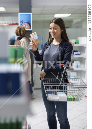 Asian customer examining medication on retail shelves during a pharmacy visit, seeking self care medical supplies from the apothecary. Female client reviewing the store merchandise. 133566106
