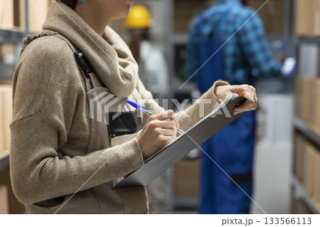 Female employee counting supplies boxes on racks for daily depot operations, focusing on goods dispatch and quality control. Pallets and boxes stacked in the industrial warehouse. Female employee counting supplies boxes on racks for daily depot operations, focusing on goods dispatch and quality control. Pallets and boxes stacked in the industrial warehouse. 133566113