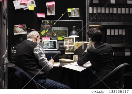 Two inspectors conducting detective work using a landline phone for inquiry and reporting in an incident room. Detectives cross checking evidence with the forensic department on the line. 133566117