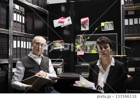 Portrait of police agents studying case files and records in evidence room, connecting documents and photos on a mystery board to uncover a suspect in organized crime. Conducting detective work. 133566118