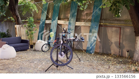 Two bicycles in backyard waiting for repair and maintenance with professional equipment as annual summer activity. Broken bike positioned on repair-stand for examining and fixing outside. 133566156