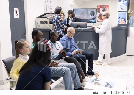 Female general practitioner with clipboard sits between elderly man and young black man in clinical lobby. Diverse patients wait nearby for medical consultations in modern hospital waiting room. 133566171