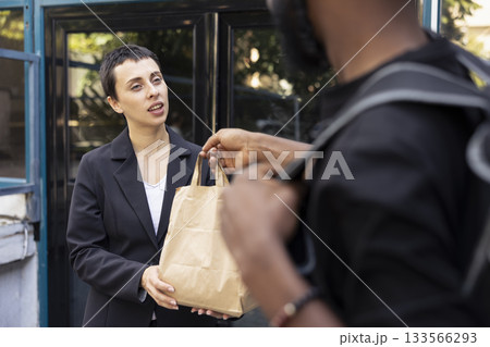 Modern delivery person arrives to deliver fast food to a woman client, using a mobile application for express restaurant delivery. Quick service takeout and urban lifestyle, paper bag. Modern delivery person arrives to deliver fast food to a woman client, using a mobile application for express restaurant delivery. Quick service takeout and urban lifestyle, paper bag. 133566293
