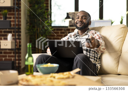 African american freelancer works remotely from sofa with digital device on his lap, using remote to adjust TV volume. Young man multitasking between laptop and watching television at home. African american freelancer works remotely from sofa with digital device on his lap, using remote to adjust TV volume. Young man multitasking between laptop and watching television at home. 133566308