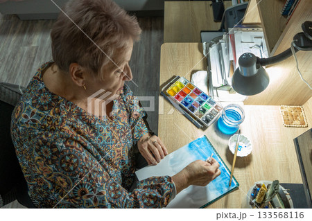 An elderly woman paints with watercolors at home. 133568116