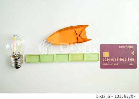 Top view of a lightbulb with rectangular blocks and a credit card with a paper sailboat on a white background. Concept of finance and travel Top view of a lightbulb with rectangular blocks and a credit card with a paper sailboat on a white background. Concept of finance and travel 133568307