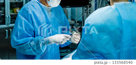 A surgeon wearing sterile gloves holds a large syringe containing medication. A surgeon wearing sterile gloves holds a large syringe containing medication. 133568540