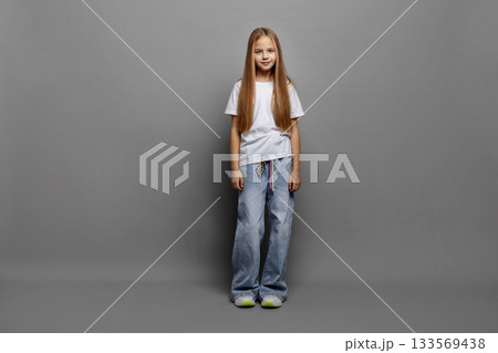 Full length portrait of beautiful girl model standing on gray studio background in white t-shirt and jeans. Cute young confident child with long blonde hair and pink highlights looking at camera 133569438
