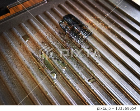 Burnt Food Residue on Old Grill Pan. Close-up of blackened, charred burnt food stuck to the ridges of a well-used metal grill pan, with grease and water droplets. 133569654