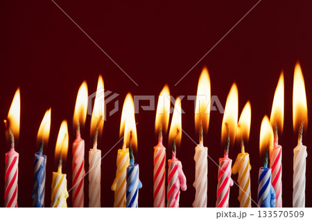 Row of colorful burning birthday candles on red background. Bright flames and vibrant colors with space for text or graphic elements. Ideal for celebration concepts. Row of colorful burning birthday candles on red background. Bright flames and vibrant colors with space for text or graphic elements. Ideal for celebration concepts. 133570509
