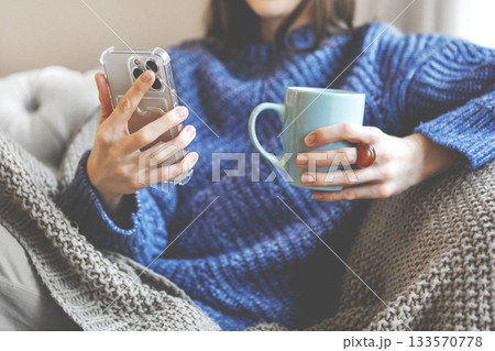 Woman shopping online at home, holding smartphone and warm cup in knitted blanket and sweater, enjoying comfort, convenience and modern digital lifestyle indoors. Cozy winter moment 133570778