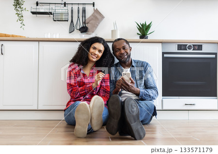 A smiling couple sits on the floor in their kitchen, using a credit card and smartphone for online shopping. 133571179