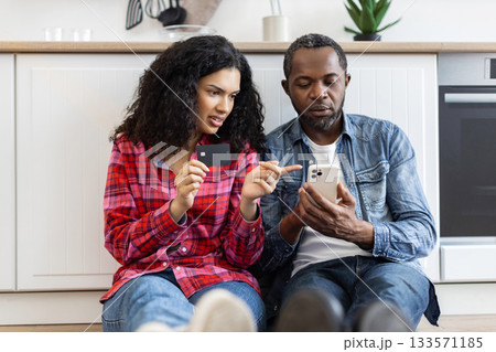A couple is sitting together, looking at a phone and a credit card in a kitchen setting. A couple is sitting together, looking at a phone and a credit card in a kitchen setting. 133571185