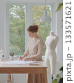 Female fashion designer drawing clothing patterns at a wooden table in a well lit studio. Creating new apparel designs with a sewing mannequin standing nearby 133571621