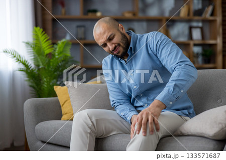 Man feeling intense pain in his knee, sitting on a gray sofa at home, touching his leg, experiencing discomfort and health issues, requiring care for joint and muscle problems Man feeling intense pain in his knee, sitting on a gray sofa at home, touching his leg, experiencing discomfort and health issues, requiring care for joint and muscle problems 133571687