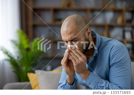 Sad man sitting on a sofa at home during cold and flu season, blowing his nose with a tissue, looking unwell and fatigued from runny nose and nasal congestion Sad man sitting on a sofa at home during cold and flu season, blowing his nose with a tissue, looking unwell and fatigued from runny nose and nasal congestion 133571693