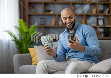 Happy young man holding cash and smartphone, celebrating online financial success and profit, receiving good news about earnings or a banking app transaction while sitting on a couch 133571696