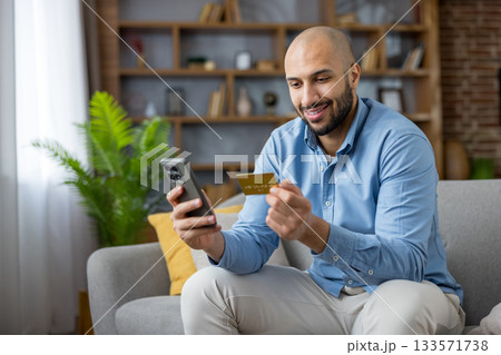 Man in a casual shirt sitting on a sofa in a modern living room, smiling and holding a smartphone and credit card, engaged in online shopping and banking 133571738
