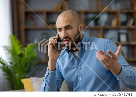 Young man on a mobile phone call receiving bad news, showing stress, worry and frustration during a tense conversation at home, conveying conflict and disappointment 133571741