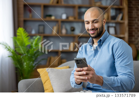Young man smiling and relaxing on a couch at home, browsing social media on his smartphone while engaging with digital content, representing modern communication and leisure Young man smiling and relaxing on a couch at home, browsing social media on his smartphone while engaging with digital content, representing modern communication and leisure 133571789