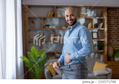 Smiling man standing in a modern living room, showing loose pants after successful weight loss, embracing a new healthy lifestyle and fitness transformation 133571790