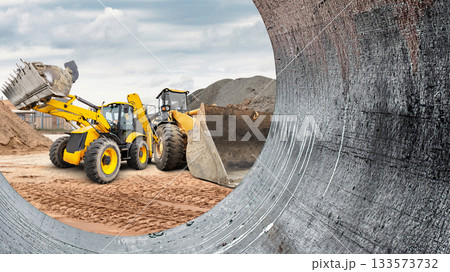 Two pieces of heavy machinery are busy moving dirt and materials at a construction site under a cloudy sky in an industrial area 133573732