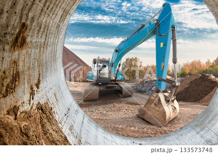 A blue excavator is digging at a construction site surrounded by dirt piles and trees under a bright sky during the day 133573748