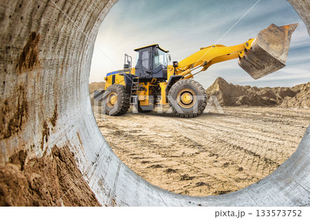 A yellow loader is seen through a concrete pipe at a construction site, with rugged mountains in the background during sunset hours. Dust rises from the ground 133573752