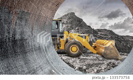 A yellow front loader moves earth at a construction site framed by a large pipe. Mountains loom in the background under an overcast sky, indicating ongoing work 133573758