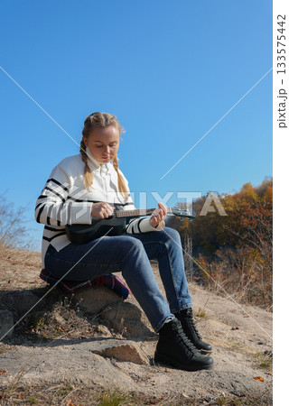 Beautiful blonde girl playing guitar ukulele and singing relaxing enjoying outdoors on sunny autumn day. Hippie woman in boho style training playing musical instrument. Fall leaves 133575442