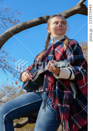 Blonde woman playing guitar ukulele outdoors in autumn forest. Concept of sound therapy, mental health and wellness rituals. Calmness tranquility audio-sensory practices. Aura farming 133575443