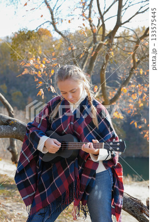 Beautiful blonde girl playing guitar ukulele and singing relaxing enjoying outdoors on sunny autumn day. Hippie woman in boho style training playing musical instrument. Fall leaves 133575445