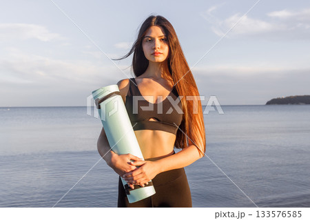 Woman holding yoga mat by the sea at sunset, fitness lifestyle, yoga training, outdoor wellness, healthy routine, seaside workout, calm evening yoga vibes. 133576585