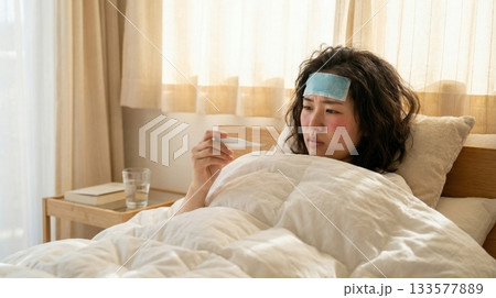 ベッドで横になり検温結果を確認する風邪の日本人女性 133577889