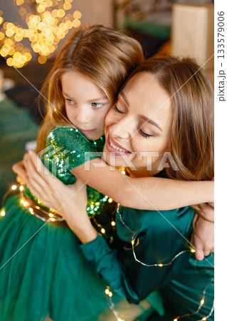 Portrait of a little girl celebrating Christmas with her mother at home. Girl and her mother posing for a photo. Kid and woman wearing green dresses and holding Christmas lights. Portrait of a little girl celebrating Christmas with her mother at home. Girl and her mother posing for a photo. Kid and woman wearing green dresses and holding Christmas lights. 133579006