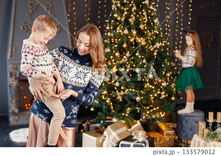 Focus on a mother holding her son on hands. Young mother, little daughter and son in studio posing for a photo. Woman and kids wearing knitted sweaters with Christmas prints. Focus on a mother holding her son on hands. Young mother, little daughter and son in studio posing for a photo. Woman and kids wearing knitted sweaters with Christmas prints. 133579012